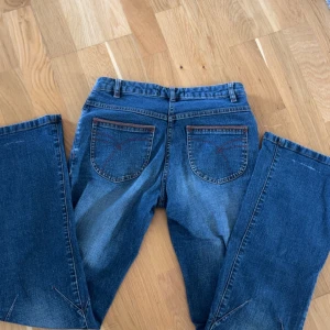 Blå bootcut jeans med flätad detalj - Snygga blå jeans från Lindex med bootcut passform. Jeansen har flätad detalj vid fickorna och kontrastsömmar i rött. Klassisk femficksmodell med dragkedja och knapp. Perfekta för dig som gillar en avslappnad men trendig look.