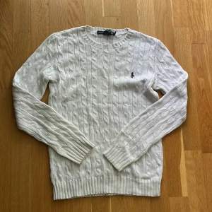 Vit kabelstickad tröja från Ralph Lauren - Vit kabelstickad tröja från Ralph Lauren med klassisk rund halsringning och broderad logga på bröstet. Tröjan har långa ärmar och ribbade muddar vid ärmslut och nederkant. Perfekt för lager-på-lager och stilren look.
