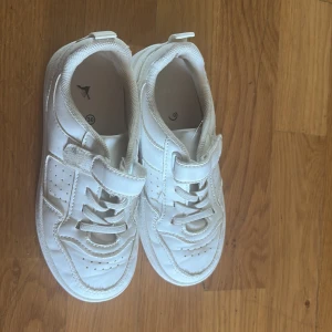 Vita sneakers med kardborreband - Snygga helvita sneakers i syntetmaterial med både snörning och praktiskt kardborreband. Skorna har perforerade detaljer på sidorna och en platt sula. Perfekta för en clean och sportig look.