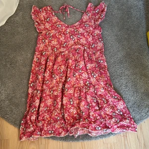 Rosa blommig sommarklänning med volang - Supersöt rosa blommig klänning med volangdetaljer och knyt i nacken. Lätt och luftig modell med kort ärm och v-ringning, perfekt för varma dagar. Färgglada blommor i vitt, gult och blått ger en härlig sommarkänsla.vet inte storlek då det inte står men skulle vilja säga s kanske m men skulle ändå säga s