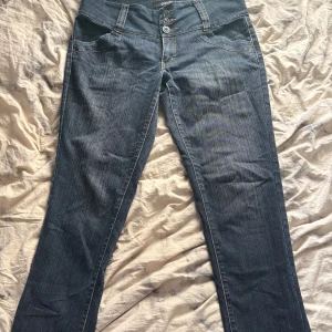 Jeans  - Snygga blå jeans från Gianini med hög midja, raka ben och coola detaljer som dragkedjor på bakfickorna och strassdekor upptill. Byxorna har tre knappar framtill och klassisk jeanslook med kontrastsömmar.
