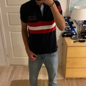 Race Marine pikétröja XS, marinblå/röd - Snygg pikétröja från Race Marine i marinblått med röda och vita ränder över bröstet. Klassisk krage med ljusblå knappslå och broderad text på bröstet. Röd siffra 8 på ärmen. Perfekt för en sportig och stilren look.