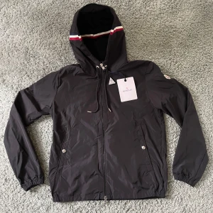 Moncler windbreaker i svart - Svart Moncler vindjacka med huva och dragsko, detaljerad med röd och vit rand på huvan. Jackan har dragkedja framtill, två fickor med tryckknappar och Moncler-logga på ärmen. Perfekt för dig som gillar stilren och sportig look. Den är helt ny och kommer med taggarna och dem är i storlek M.