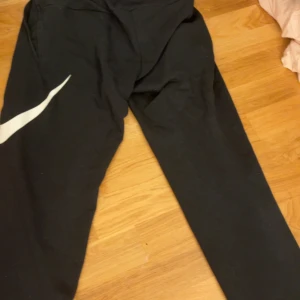 Svarta Nike Dri-Fit mjukisbyxor - Svarta mjukisbyxor från Nike med Dri-Fit-teknologi. Byxorna har elastisk midja med Nike-logga, snedställda fickor och en stor vit swoosh på ena benet. Perfekta för träning eller chill, med skön passform och sportig look. Storlek S använt 2 gånger