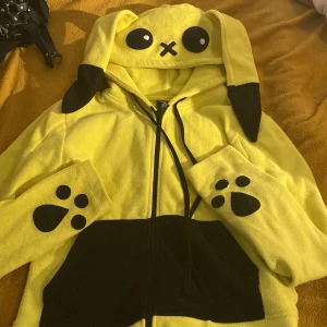 Gul fleecejacka med Pokémon - Superunik gul fleecejacka från capcake cult med svarta detaljer, stora kaninöron på luvan och broderade ögon. Svarta tassavtryck på ärmarna och blixtmönster på ryggen. Dragkedja framtill och mjukt fleece-material. Perfekt för dig som gillar statement-plagg!