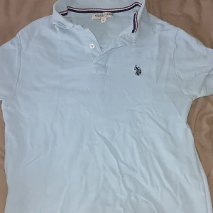 Ljusblå pikétröja från U.S. Polo Assn. - Säljer en ljusblå pikétröja från U.S. Polo Assn. med klassisk krage och knappslå. Tröjan har broderad logga på bröstet och en snygg randig detalj på insidan av kragen. Perfekt för en clean och sportig stil.