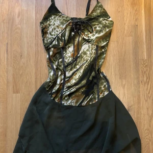 Grön camo-inspirerad kortklänning - Snygg kortklänning med camo-inspirerat mönster i grönt, brunt och svart. Klänningen har smala axelband, snörning framtill och en flowy kjol i tunnare tyg