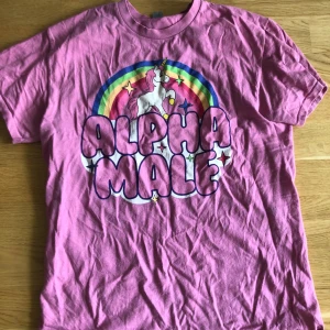 Rosa t-shirt med enhörning och regnbåge - Rosa t-shirt med coolt tryck av en enhörning, regnbåge och texten 'Alpha Male' i stora bubbelbokstäver. Färgglada detaljer och stjärnor gör den extra unik. Perfekt för dig som gillar statement-plagg och lekfull stil.