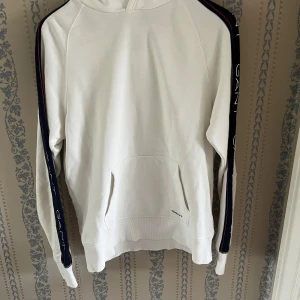 Vit hoodie från GANT med logga på ärmar - Vit hoodie från GANT med huva och känguruficka. Ärmarna har marinblå band med GANT-logga och röda detaljer längs sidorna. Perfekt för en chill och sportig look. Klassisk passform och mjukt material.