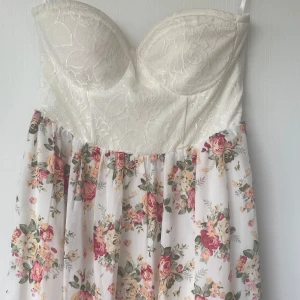 Vit långklänning med spets och blommor - Romantisk långklänning i vitt med axelbandslös överdel i spets och vadderade kupor. Kjolen är i ett lätt, flowy tyg med blommigt mönster i rosa, gult och grönt. Dragkedja bak för enkel påklädning. Perfekt för sommarens alla tillfällen!