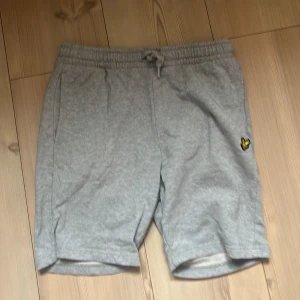 Grå mjukisshorts Lyle & Scott  - Grå mjukisshorts från Lyle & Scott Junior med elastisk midja och snörning. Klassisk gul logga broderad på ena benet. Shortsen har sidofickor och en bakficka, perfekta för chill dagar eller träning.