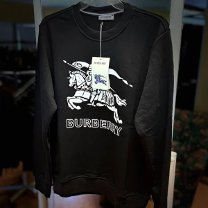 Svart Burberry sweatshirt med tryck - Svart sweatshirt från Burberry i 100% bomull med stort vitt tryck av riddare och logga på bröstet. Klassisk rund halsringning och långa ärmar. Perfekt för dig som vill ha en stilren och lyxig streetwear-look.