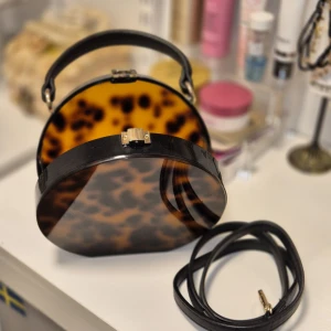 Rund leopardmönstrad handväska - Säljer en unik rund handväska med leopardmönster i brunt och svart. Väskan har ett hårt skal, svart handtag och medföljande axelrem. Stängs med ett guldfärgat spänne och har cool retrovibe. Perfekt för dig som vill sticka ut med accessoarerna.