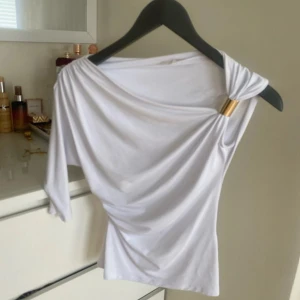 Vit topp med gulddetalj från Zara - Supersnygg vit topp från Zara med en axel och guldfärgad metallring som cool detalj. Toppens tyg faller snyggt och har en lätt drapering. Perfekt för dig som vill sticka ut med en unik och trendig look.