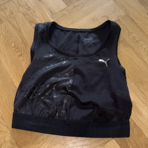 Svart sportig linnetopp från Puma - Svart linnetopp från Puma med glansiga detaljer och brett, texturerat band nedtill. Toppen är ärmlös och har en sportig look med diskret Puma-logga framtill. Perfekt för dig som gillar att träna och vill ha en snygg och bekväm topp.