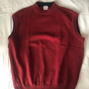 Vinröd stickad väst från Boomerang - Snygg vinröd stickad väst från Boomerang Sportswear med broderad logga på bröstet. Ribbade kanter vid hals, ärmhål och nederkant. Perfekt att slänga över en skjorta eller tshirt. Storlek L.