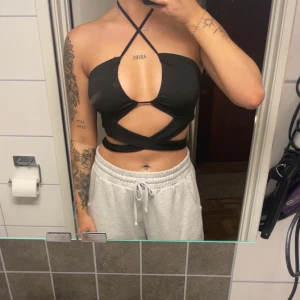 Svart omlott croppad tubtopp - Säljer en svart croppad tubtopp med omlott och snörning som knyts både i nacken och runt midjan. Toppen har en djup v-ringning framtill och är axelbandslös med coola cut-outs. Perfekt för dig som vill sticka ut med en trendig och edgy look.