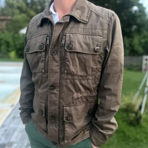 J.Lindeberg feild jacka - Snygg olivgrön jacka från J.Lindeberg i storlek M. Jackan har fyra stora fickor framtill med knappar, hög krage och både dragkedja och knäppning. Perfekt för dig som gillar stilren och funktionell design. Perfekt inför hösten 👌Lite sliten längst ner vid midjan.