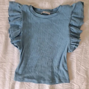 Blå ribbad topp med volangärmar - Supersöt ljusblå ribbad topp från Zara med volangdetaljer på axlarna. Toppen är ärmlös med breda volanger som ger en unik och trendig look. 