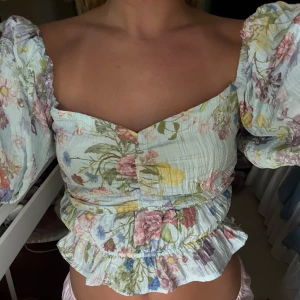 Blommig blus med puffärmar - Supersöt blommig blus i ljusa pastellfärger med volangkant och puffiga korta ärmar. Toppen har en smickrande v-ringning och smockad midja som ger en feminin siluett. Perfekt för dig som gillar romantisk och trendig stil.
