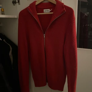 Röd stickad kofta från East West - Säljer en röd stickad kofta från East West med hel dragkedja framtill och hög krage. Tröjan har lång ärm och en clean, enkel design som passar perfekt till lager-på-lager-stil. Grym för dig som gillar färg och vill sticka ut lite extra.
