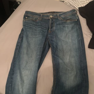 Blå jeans från Jack & Jones - Säljer ett par klassiska blå jeans från Jack & Jones med snygga slitningar och kontrastsömmar. Jeansen har fem fickor, normal passform och är tillverkade i bomull. Perfekta för en avslappnad stil.