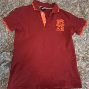 Röd pikétröja från Hugo Boss - Snygg röd pikétröja från Hugo Boss med orange detaljer på krage och ärmslut. Klassisk pikéknäppning med två knappar och tryckt logga på bröstet. Har några märken som ni ser i bilden så säljer den för låg pris.