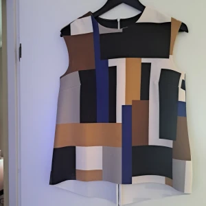 Ärmlös grafisk blus i blockmönster - Säljer en ärmlös blus med grafiskt blockmönster i svart, brun, blå, beige och grå. Toppen har rak passform och rund halsringning. Materialet är slätt och känns lite strukturerat, perfekt för dig som gillar en modern och stilren look.