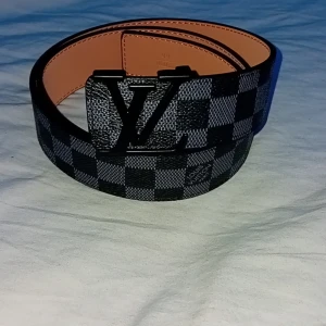 rutigt skärp med spänne - Snyggt bälte i svart och grått med klassiskt rutigt mönster och stort spänne i i svart. Bältet är gjort i skinn och har en exklusiv känsla. Perfekt accessoar för att lyfta din outfit med en lyxig touch.