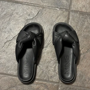 Svarta sandaler från MNG med knut - Stilrena svarta sandaler från mango med breda remmar i skinnimitation och snygg knutdetalj upptill. Platt sula och öppen tå gör dem perfekta för varma dagar. Enkla att matcha till olika outfits. Knappt använda.