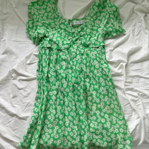 Grön blommig kortklänning från Zara - Supersöt grön kortklänning från Zara med vita och rosa blommor. Klänningen har korta ärmar, v-ringning och knytning framtill. Perfekt för varma dagar och ger en fräsch sommarkänsla. Lätt och luftig modell som är riktigt bekväm.