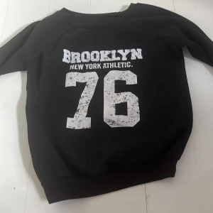 Svart sweatshirt Brooklyn 76 - Svart sweatshirt med tryck i vitt framtill: 'BROOKLYN NEW YORK ATHLETIC 76'. Tröjan har rund halsringning och långa ärmar. Perfekt för en sportig och avslappnad stil.