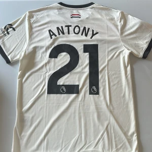 Manchester United Antony 21 bortatröja - Snygg Manchester United bortatröja med Antony 21-tryck på ryggen. Tröjan är vit med svarta och röda detaljer, svarta Adidas-ränder på axlarna och svarta muddar. Snapdragonsponsor på bröstet och Premier League-logga på ärmen. Tillverkad i lätt och ventilerande polyester. Replika. Endast handtvätt gäller.