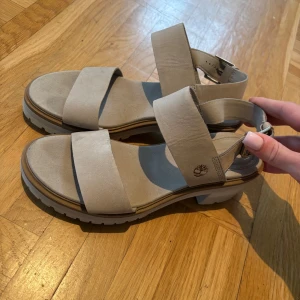 Beige sandaler från Timberland - Chunky sandaler i beige mocka från Timberland med breda remmar och justerbart spänne vid ankeln. Grov sula med låg klack och öppen tå ger en cool och modern vibe. Perfekta för varma dagar när du vill ha både stil och komfort.