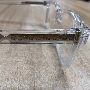 Transparenta glasögon med gulddetaljer - Snygga glasögon med transparent båge och fyrkantig form. Skalmarna har inlägg i guld med graverade detaljer som ger en unik och modern look. Perfekta för dig som vill sticka ut med stilrena accessoarer.