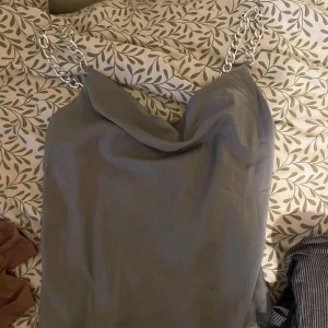 Grå linnetopp med kedjeband - Supersnygg grå linnetopp med silvriga kedjor som axelband och draperad urringning. Perfekt för dig som vill sticka ut lite extra med coola detaljer. Toppens material känns slätt och har ett lätt glansigt utseende.