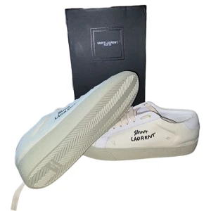 Saint Laurent vita sneakers i canvas - Saint Laurent sneakers i vit canvas med handskriven logga på sidan. Skorna har vita snören, detaljer i vitt skinn och en platt, ljus sula. Snygg minimalistisk design med en cool streetkänsla. Kommer med originalkartong och dustbag.