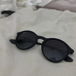 Svarta runda solglasögon Ray-Ban - Snygga svarta solglasögon från Ray-Ban med rund form och mattsvart båge. Klassisk design med mörka glas och diskret logga i hörnet. Perfekta för att lyfta din stil och skydda ögonen mot solen.
