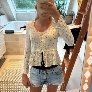 Prickig blus med knyt från Monki - Supersöt blus från Monki i beige med svarta prickar. V-ringad med knappar framtill, volang nertill och knyt i midjan. Långärmad. Perfekt att styla med jeans eller kjol för en chill look. Använd få gånger