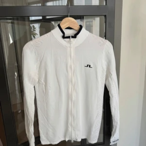 Vit zip-kofta från J.Lindeberg - Vit långärmad kofta från J.Lindeberg med hel dragkedja och svart logga på bröstet. Snygga svarta detaljer på krage och ärmslut. Lätt stickat material med diskret mönster i tyget. Super fin och endast använd 2 gånger. Perfekt på golfbanan eller på promenaden! 