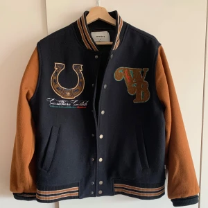 Svart och brun bomberjacka med broderier - Snygg mörkblå/brun jacka från WoodBird med broderider på bröstet och ryggen. Jackan känns väldigt premium och har snygg fit. Storlek M (jag är 182). Originalpris runt 1700kr, knappt använd. Pris går att diskuteras, hämtas i Hornstull! 🤠