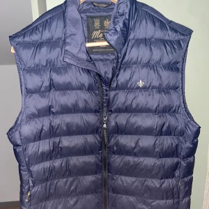 Mörkblå dunväst från Morris - Säljer en snygg mörkblå dunväst från Morris. Västen har en quiltad design och är utrustad med en dragkedja framtill. Perfekt för höst och vår när du vill hålla dig varm men ändå se stilren ut. Den är i nyskick och redo för nya äventyr!