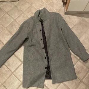 Grå rock med knappar - Säljer en stilren grå rock med svarta knappar. Den har en klassisk design med lång ärm och en snygg krage. Insidan är fodrad med ett mörkt tyg som ger en lyxig känsla. Perfekt för höst och vår! Använd ca 2 gånger 