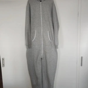 Grå onesie från Skill - Säljer en supermysig grå onesie från Skill. Den är perfekt för att chilla hemma eller ha på sig under kalla dagar. Onesien har en dragkedja framtill och en stor huva för extra komfort. Den är långärmad och har praktiska fickor på sidorna. Materialet är mjukt och skönt, perfekt för höst och vinter!