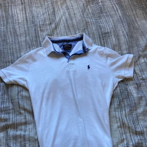 Vit pikétröja från Ralph Lauren - Säljer en klassisk vit pikétröja från Ralph Lauren i storlek S. Tröjan har en liten broderad logga på bröstet och en stilren krage med blå detaljer på insidan. 