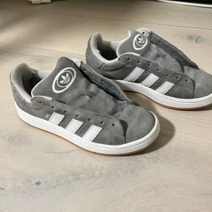 Grå Adidas campus sneakers - SNÖREN GÅR MED I KÖPET. Har haft skorna i ett år. Säljer ett par grå Adidas campus sneakers med vita detaljer och den klassiska trefoil-loggan. Skorna har en stilren design med tre vita ränder på sidan och en bekväm innersula. Perfekta för en avslappnad stil.