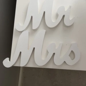 Vita bokstäver 'Mr & Mrs' - Dekorativa vita bokstäver i trä som bildar orden 'Mr & Mrs'. Perfekta för bröllop eller heminredning. Stilren design med en mjuk, rundad form.