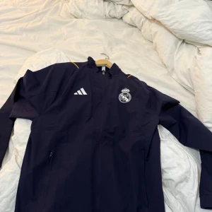 Svart träningsjacka från Adidas - Säljer en svart träningsjacka från Adidas med Real Madrid-logga på bröstet. Jackan har dragkedja och långa ärmar, perfekt för sportiga aktiviteter. Den har även vita detaljer och en gul rand på axlarna.