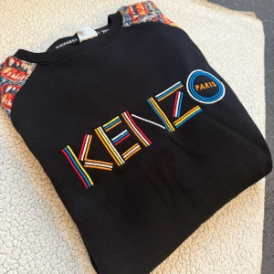 Svart tröja från Kenzo med färgglada detaljer - Säljer en svart tröja från Kenzo med färgglada broderade detaljer på ärmarna och logotypen framtill. Tröjan har långa ärmar och en unik design som sticker ut. Perfekt för dig som vill ha något annorlunda i garderoben!