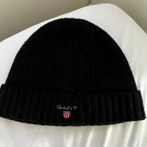 Svart ribbad mössa från GANT - Snygg svart ribbad mössa från GANT med broderad logotyp framtill. Perfekt för kyliga dagar och ger en stilren look. Mössan är tillverkad i ett mjukt material som håller dig varm och bekväm.